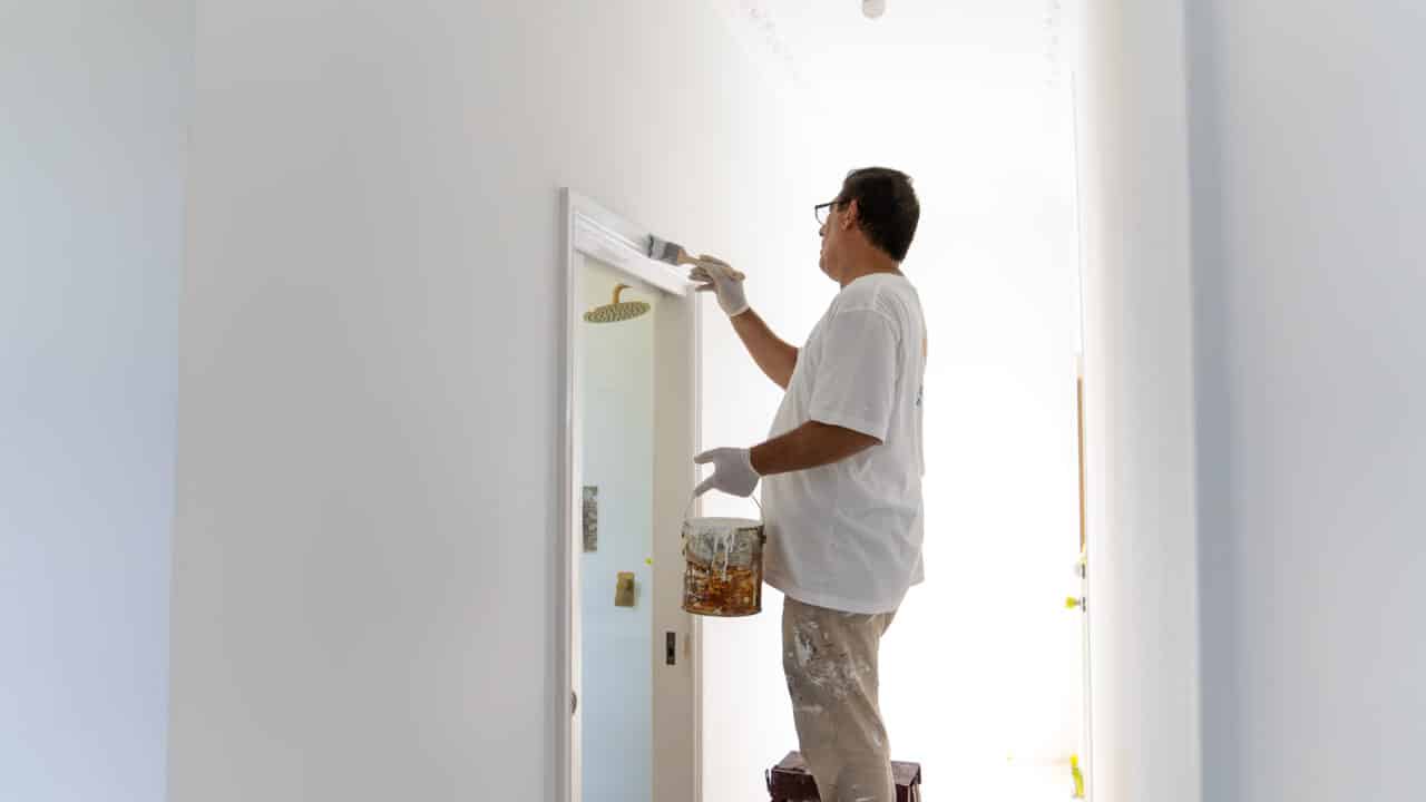 A person wearing a white shirt and gloves standing on a stool, painting a door frame in white