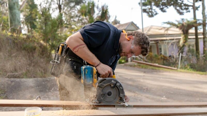 Person using a circular saw to cut wood outdoors, wearing safety glasses and a tool belt