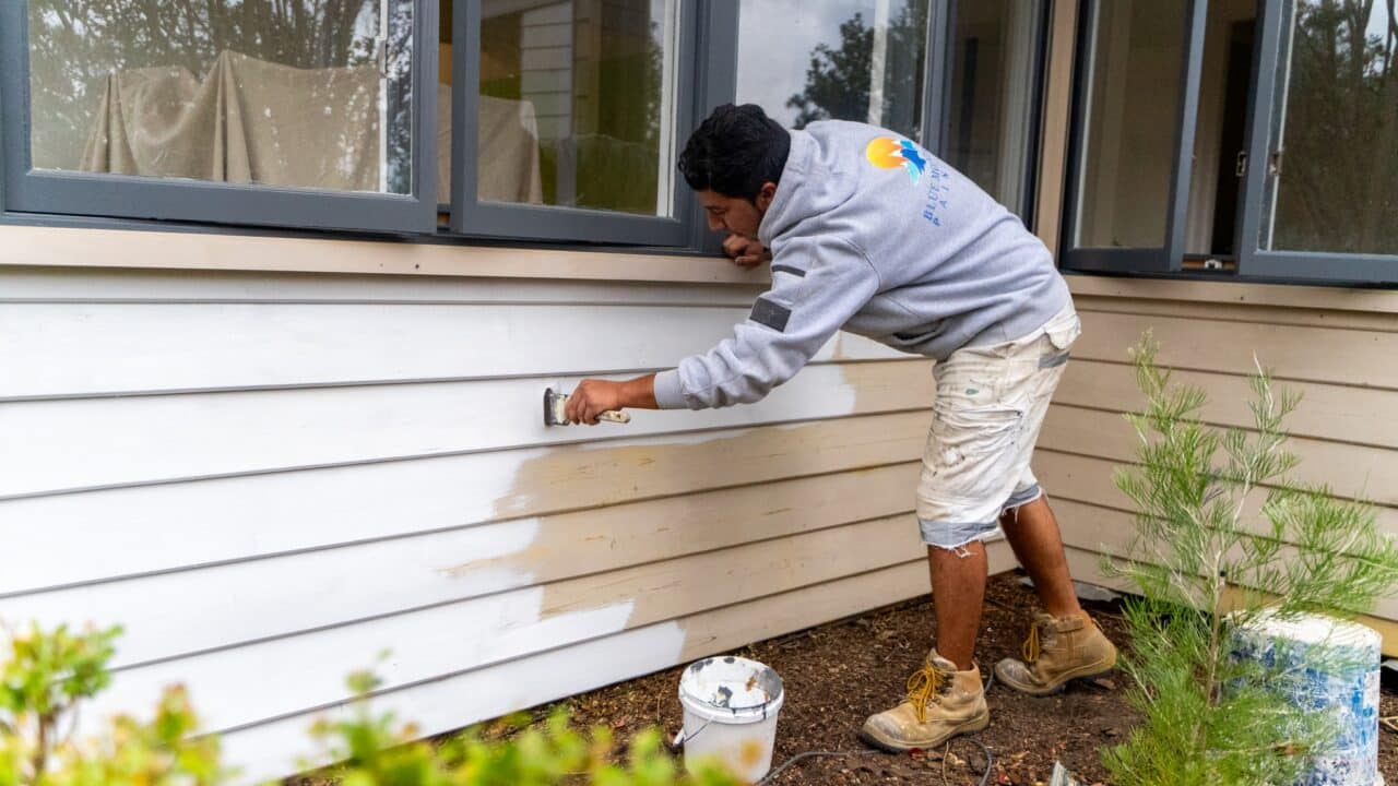 A man painting the exterior of a house white, wearing a grey hoodie and beige shorts.