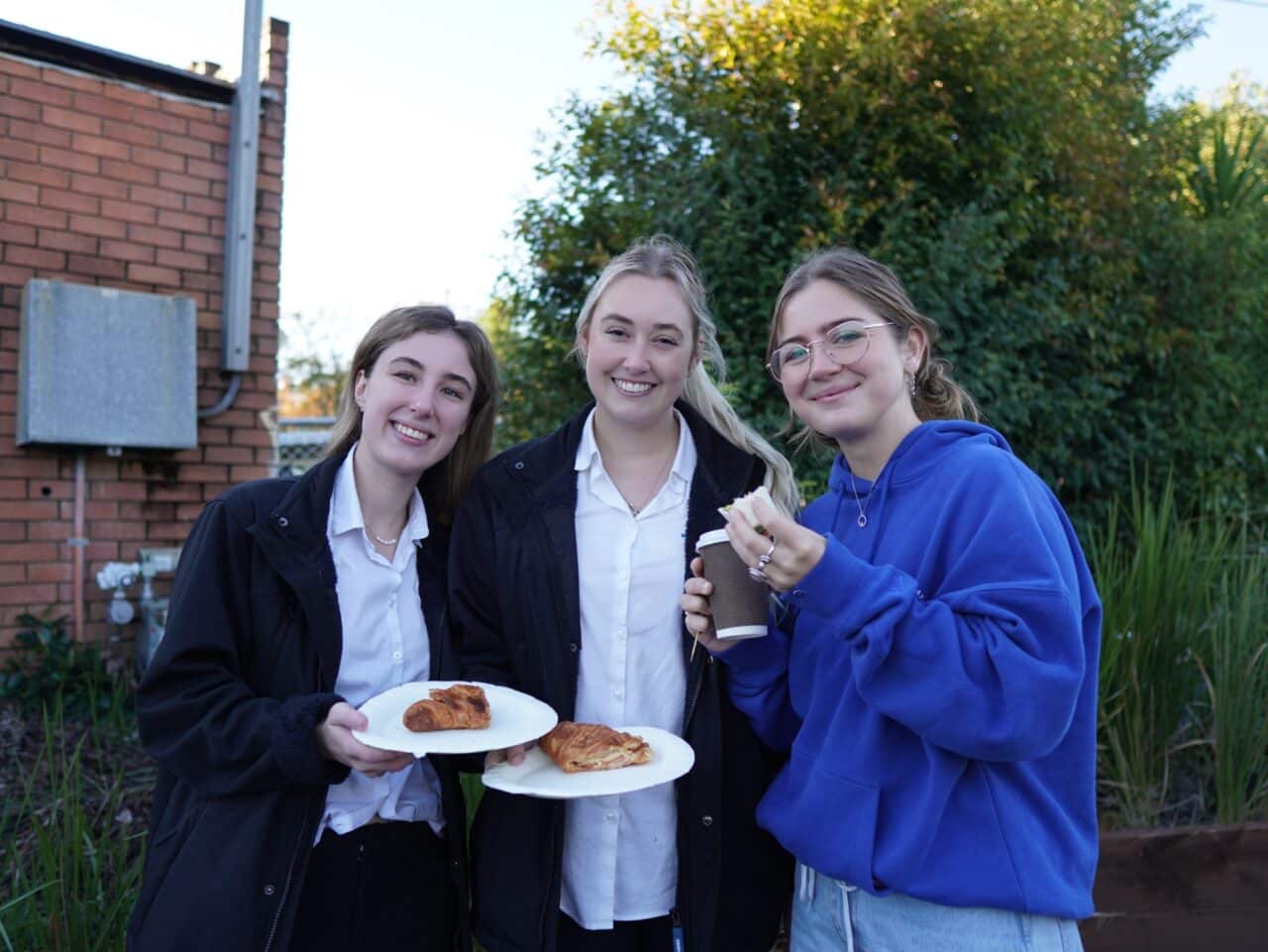 Three people standing outside; two hold plates with croissants, and one holding a cup