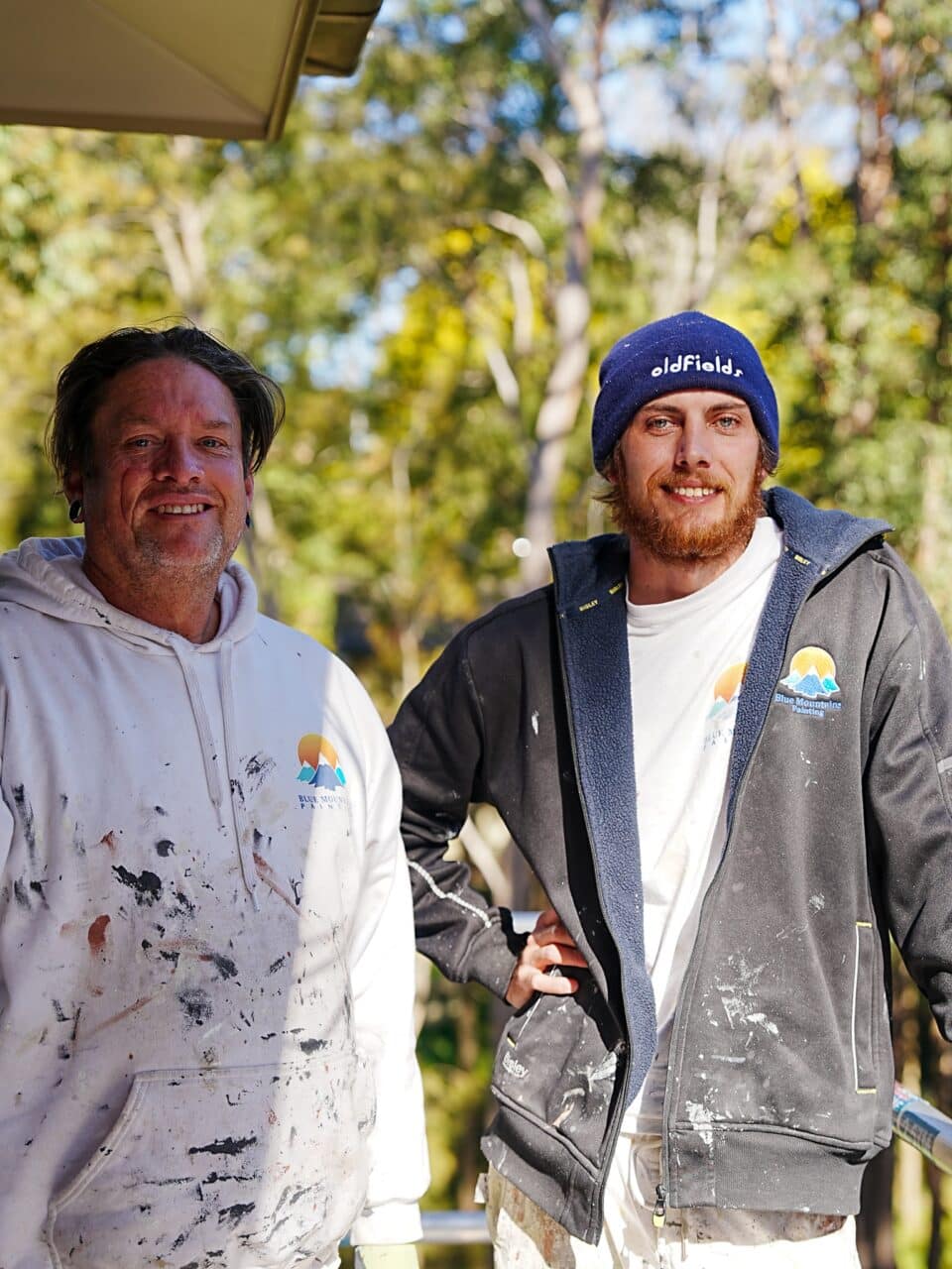 Two men in paint-splattered clothing stand outdoors, smiling