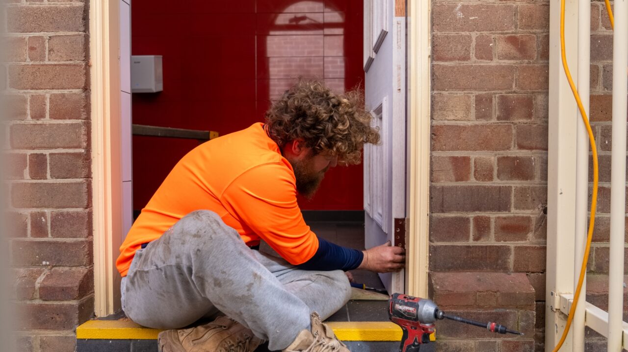 Man wearing an orange shirt and gray pants seated while installing a door frame in a brick-wall