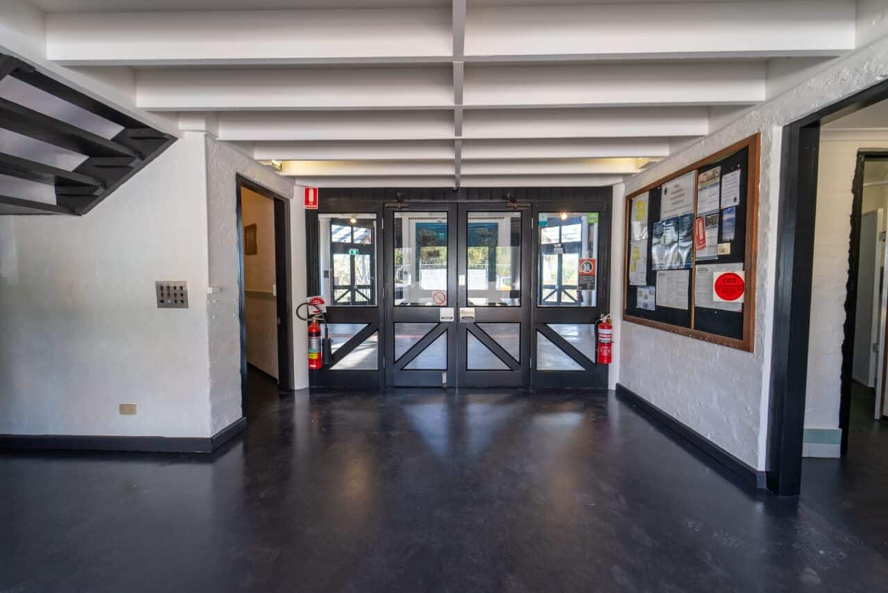 A hallway with a dark floor, white walls, a staircase on the left, and a glass double exit door.