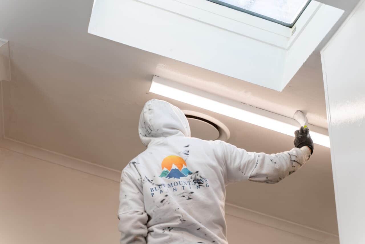 A person in a white hoodie with paint splatters painting a ceiling near a fluorescent light.