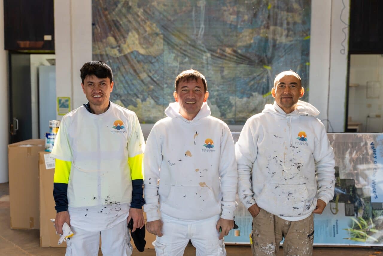 Three men in paint-splattered clothing stand in front of a large map inside a hut.