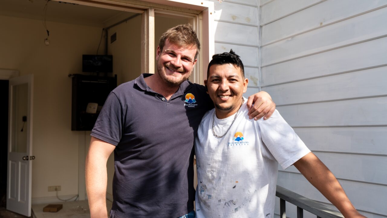 Two men smiling with arms around each other, standing outside a white building.