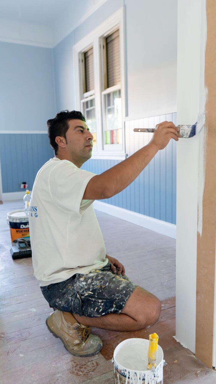 A man wearing a white T-shirt and denim shorts, kneeling on the floor, and painting a white wall