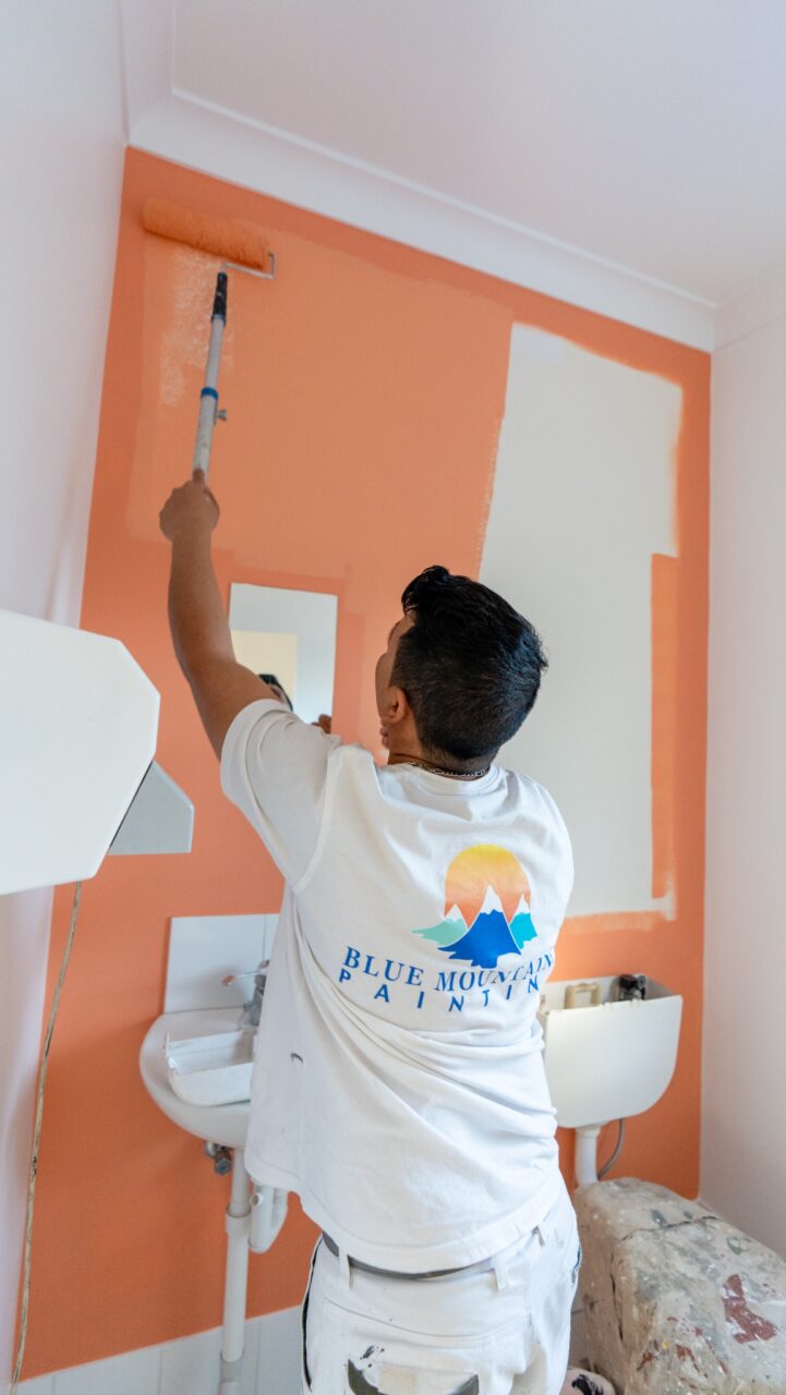 Person in a white T-shirt with a "Blue Mountain Painters" logo painting a wall orange