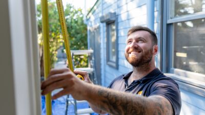 Man in a polo shirt with tattoos on his arms measures a wall outdoors using a measuring tape