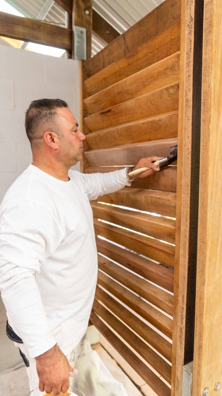 A person in a white shirt and paint-stained pants is applying a dark stain to a wooden fence