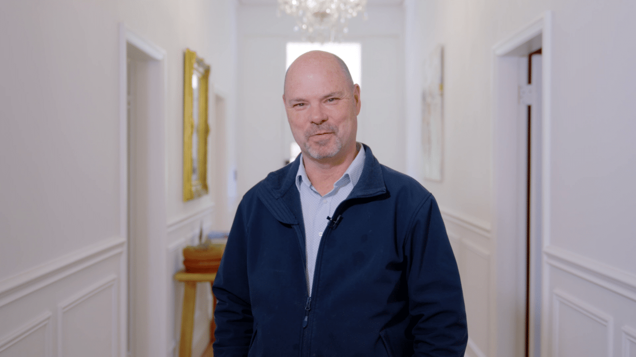 A man with a bald head and a short beard, wearing a blue jacket and smiling